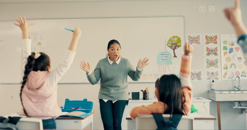 Guru wanita memandu murid-murid yang antusias mengacungkan tangan dalam kegiatan belajar interaktif di kelas