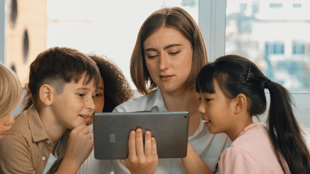 Seorang guru wanita memegang tablet sambil menjelaskan materi secara langsung kepada murid-muridnya