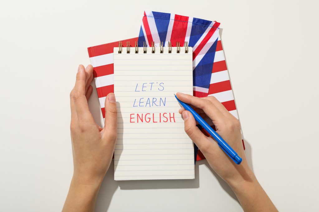angan seseorang memegang pulpen dan buku catatan bertuliskan Lets Learn English untuk persiapan tes bahasa Inggris