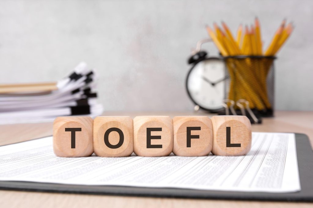 Balok kayu membentuk tulisan TOEFL yang tertata di atas kertas ujian dan meja belajar
