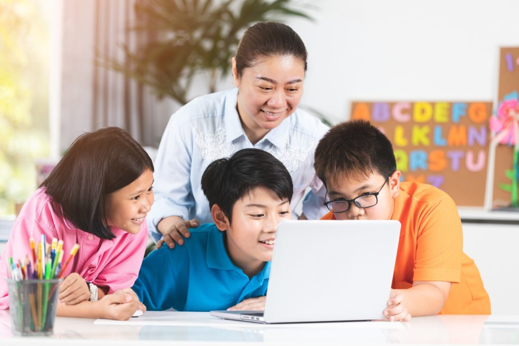Pengajar mendampingi siswa menggunakan laptop bersama dalam kegiatan belajar di kelas.